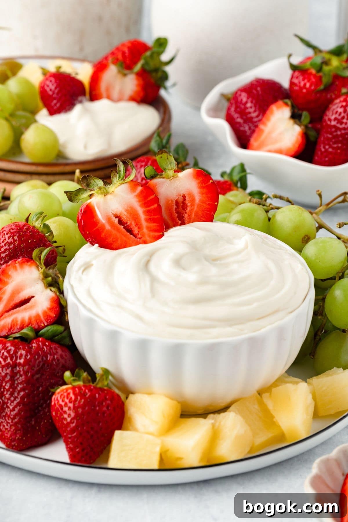 A white bowl filled with Marshmallow Fruit Dip is surrounded by fresh strawberries, green grapes, and pineapple chunks on a platter. More strawberries and grapes are in dishes in the background.