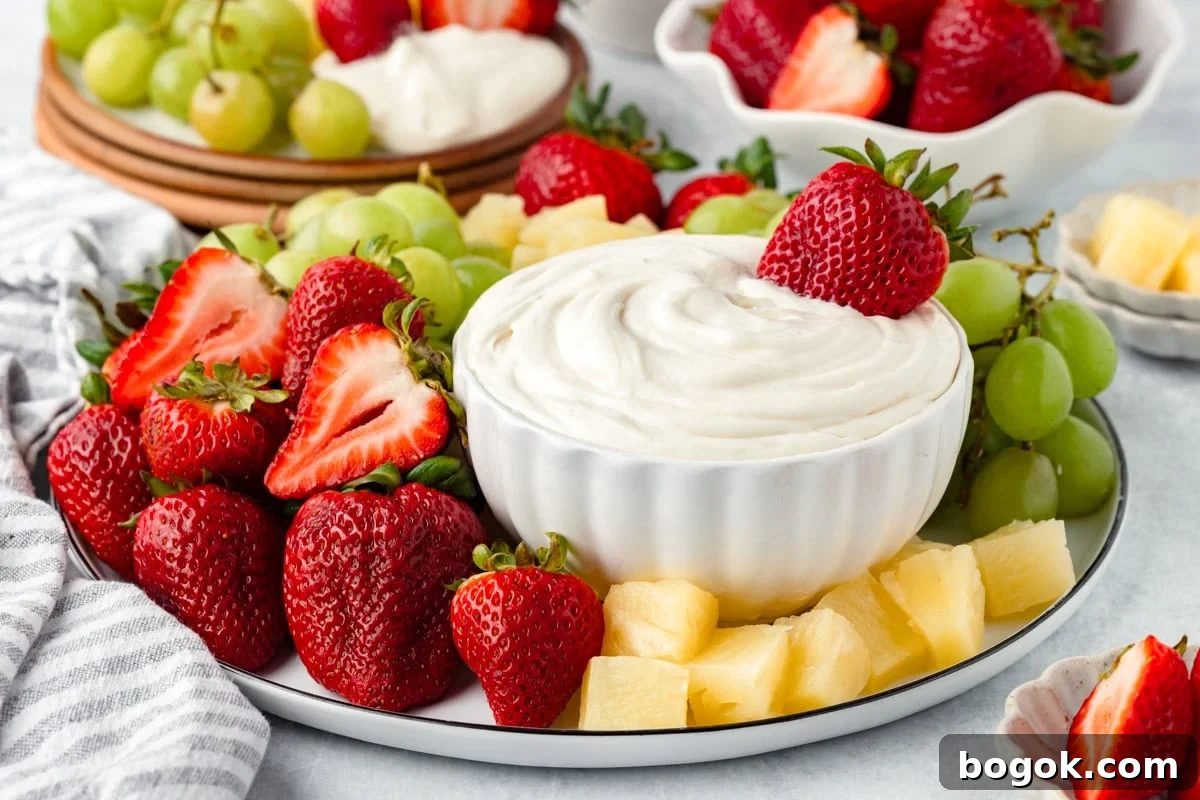 A plate with fresh strawberries, green grapes, pineapple chunks, and a bowl of creamy marshmallow fruit dip in the center, surrounded by more fruit and dip in the background.