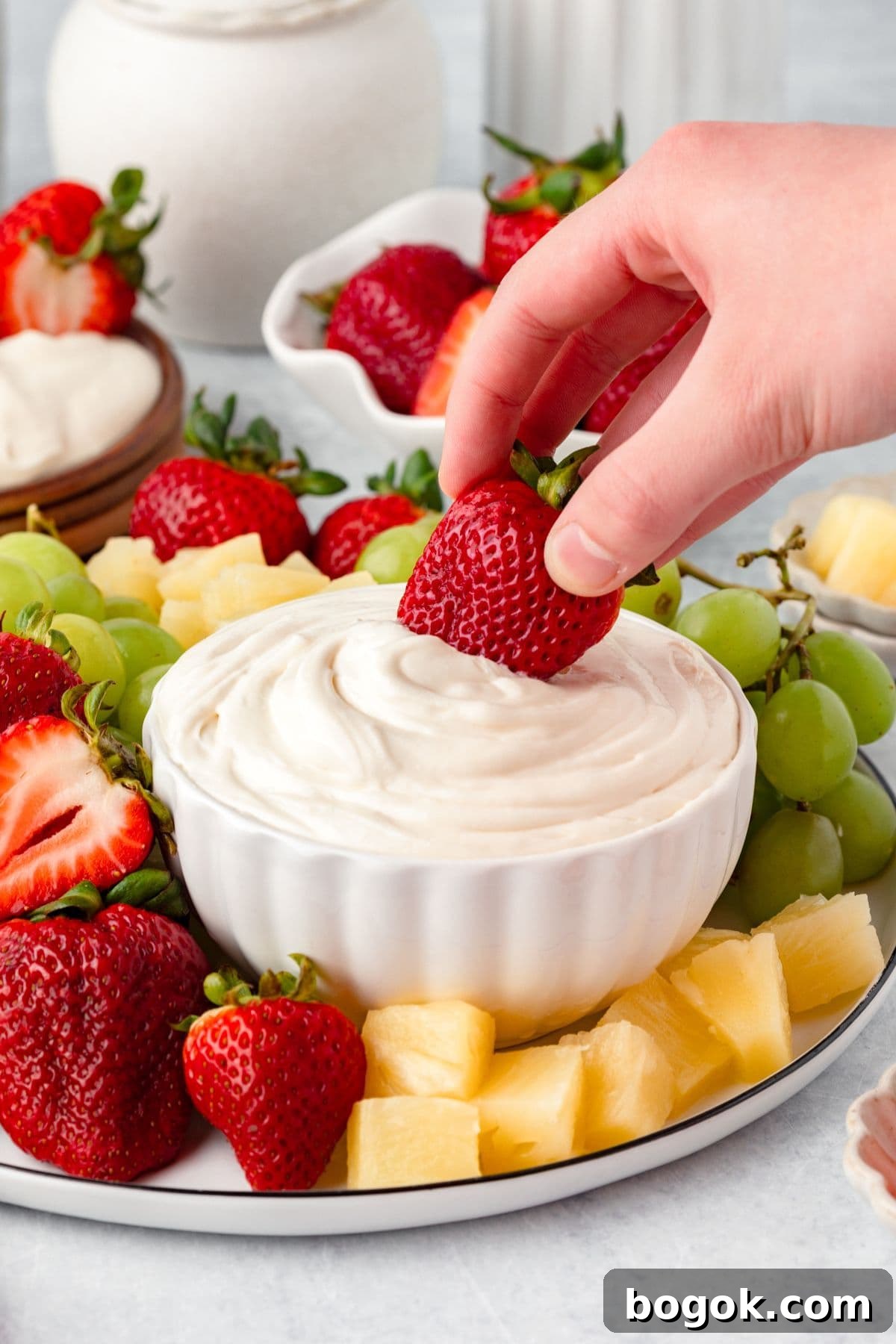 A hand dips a fresh strawberry into a bowl of Marshmallow Fruit Dip, surrounded by grapes, pineapple chunks, and more strawberries on a white platter.