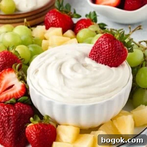 A white bowl of creamy Marshmallow Fruit Dip is surrounded by fresh strawberries, green grapes, and pineapple chunks. One whole strawberry is dipped in the luscious bowl.