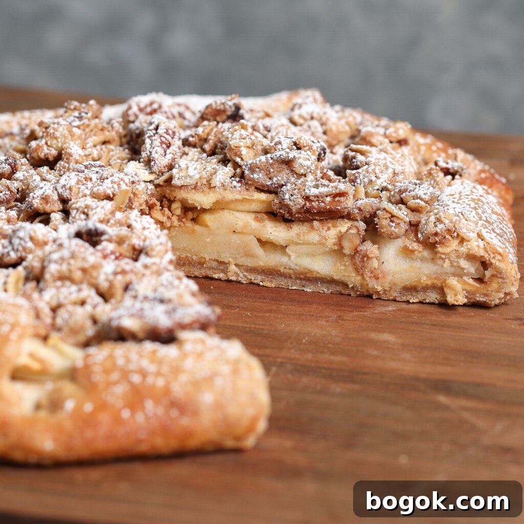 A rustic Apple Crisp Galette with Cashew Butter features a crumbly topping and powdered sugar, sliced on a wooden surface to reveal layers of tender apple filling inside.