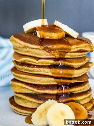 A stack of almond flour banana pancakes with syrup being poured on them and banana slices on top of them.