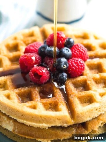 A stack of paleo waffles with berries on top and syrup being poured on them.
