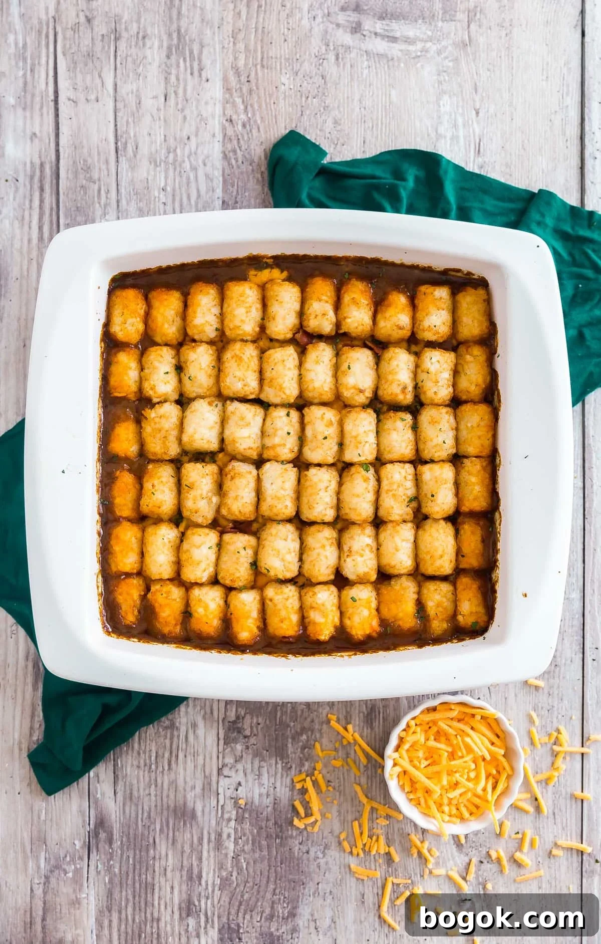 Bacon Cheeseburger Tater Tot Casserole Recipe 3 An overhead image of a cheeseburger casserole covered in tater tots on a green towel with shredded cheddar cheese next to it.