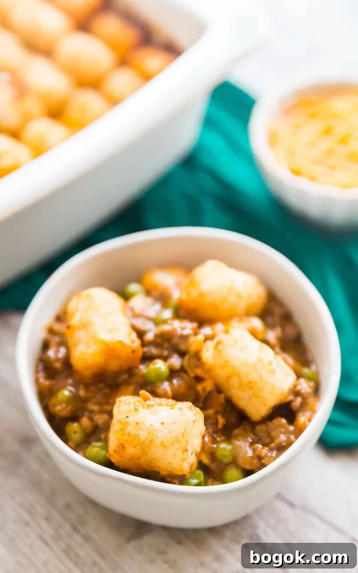Bacon Cheeseburger Tater Tot Casserole Recipe 10 A bowl of tater tot casserole with the pan behind it, a green towel and shredded cheese in the background.