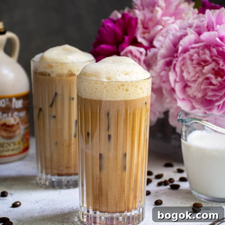 A glass filled with ice and salted maple cold brew coffee sitting on a table top next to another drink and a vase of pink flowers.