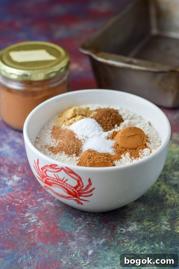 Cinnamon, ginger, cloves and nutmeg in a bowl of flour with a big jar of cinnamon in the background and a bread pan