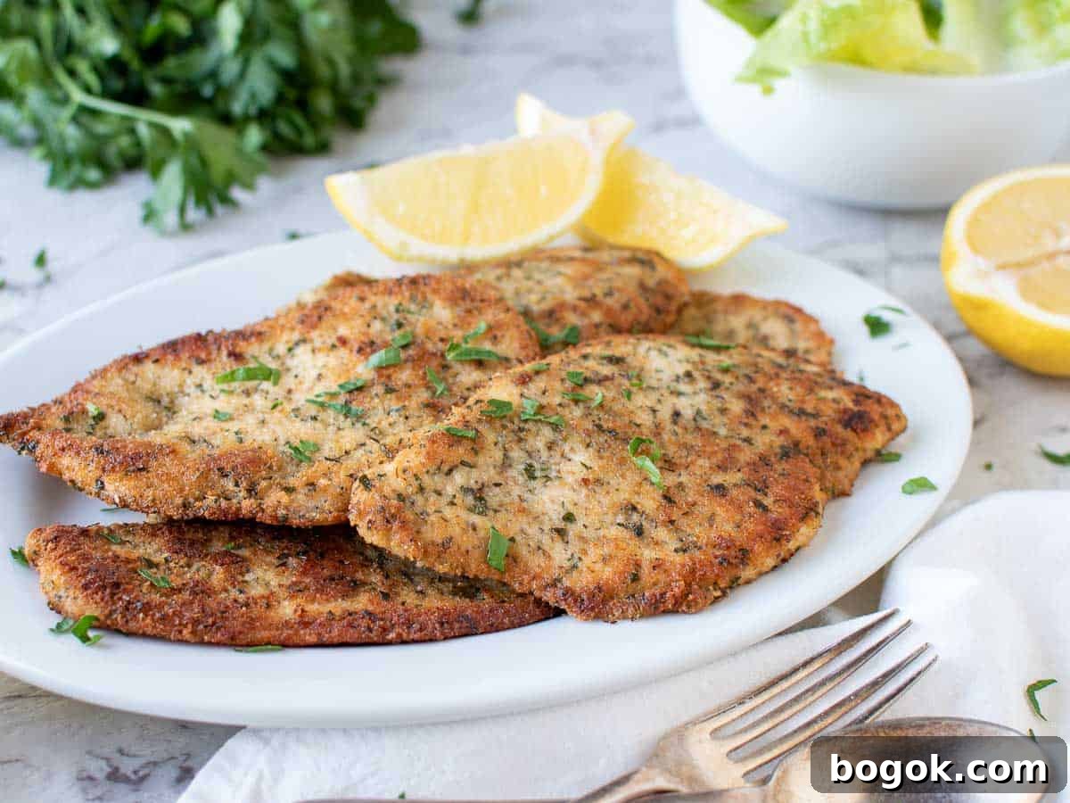 Browned chicken cutlets on white plate with lemon wedges, parsley and salad in the background.