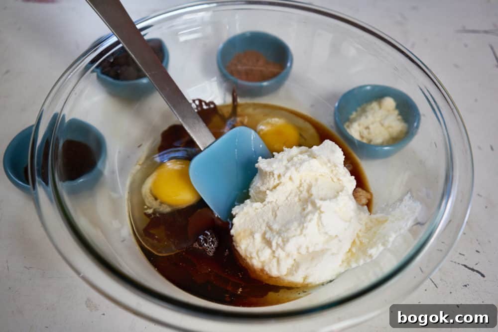 A glass bowl with a blue spatula containing ricotta cheese, eggs, vanilla and maple syrup.