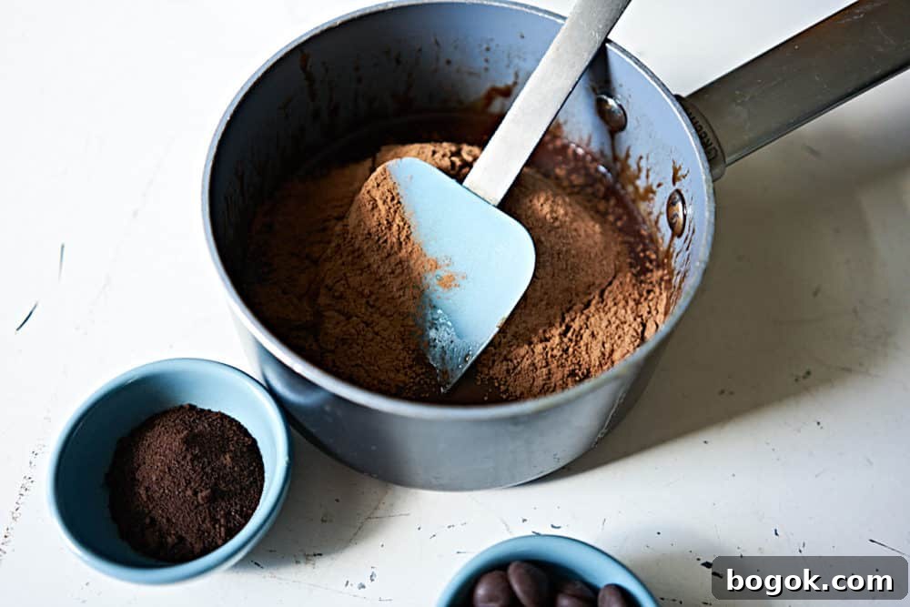A small sauce pan holds chocolate ganache, cocoa powder, and a blue spatula.