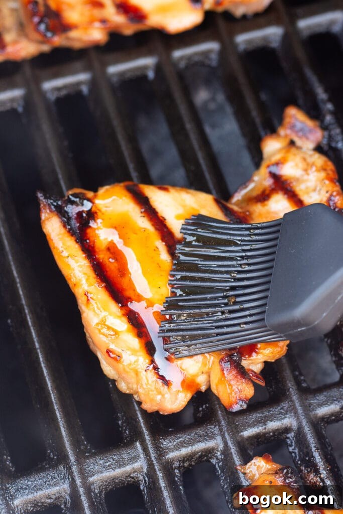 Close up of a black basting brush swiping a honey soy glaze on top of grilling boneless skinless chicken thighs on a grill.