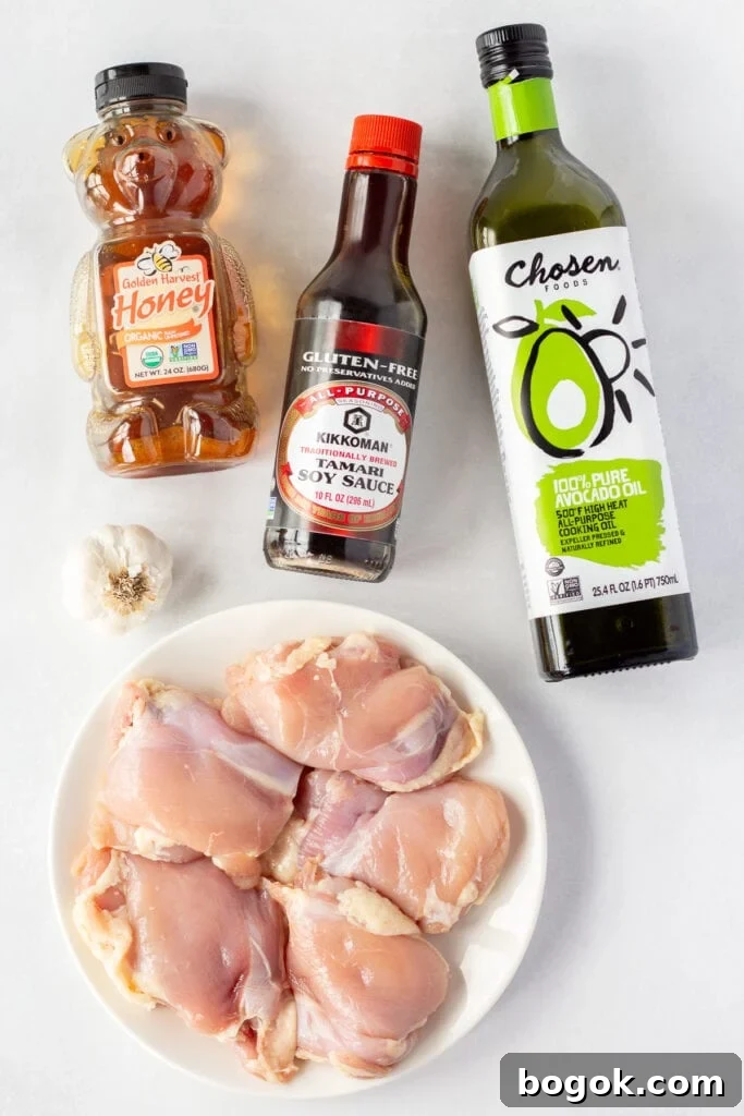 Top down shot of ingredients for grilled soy garlic chicken, including bottles of avocado oil, soy sauce, and honey, a clove of garlic, and a white plate with raw boneless skinless chicken thighs on it.