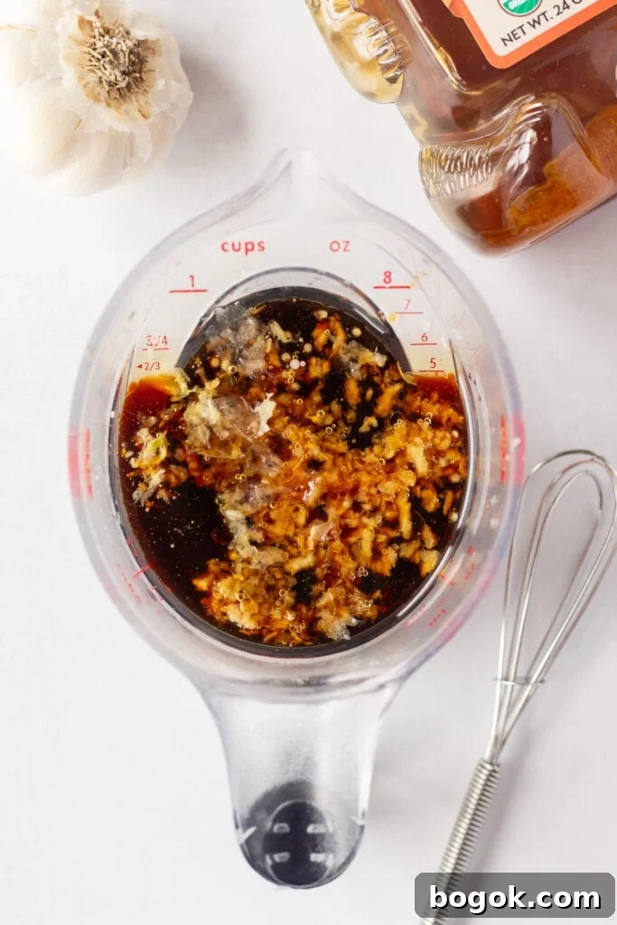 Top down shot of a measuring cup with soy sauce and garlic in it, on a white surface. Next to it are a head of garlic, a bear shaped bottle of honey, and a small whisk.