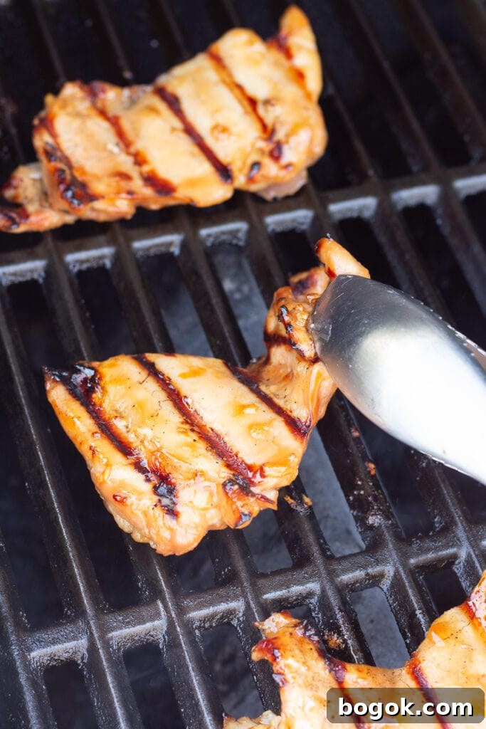 Close up of a pair of silver tongs flipping over boneless skinless chicken thighs on a grill.