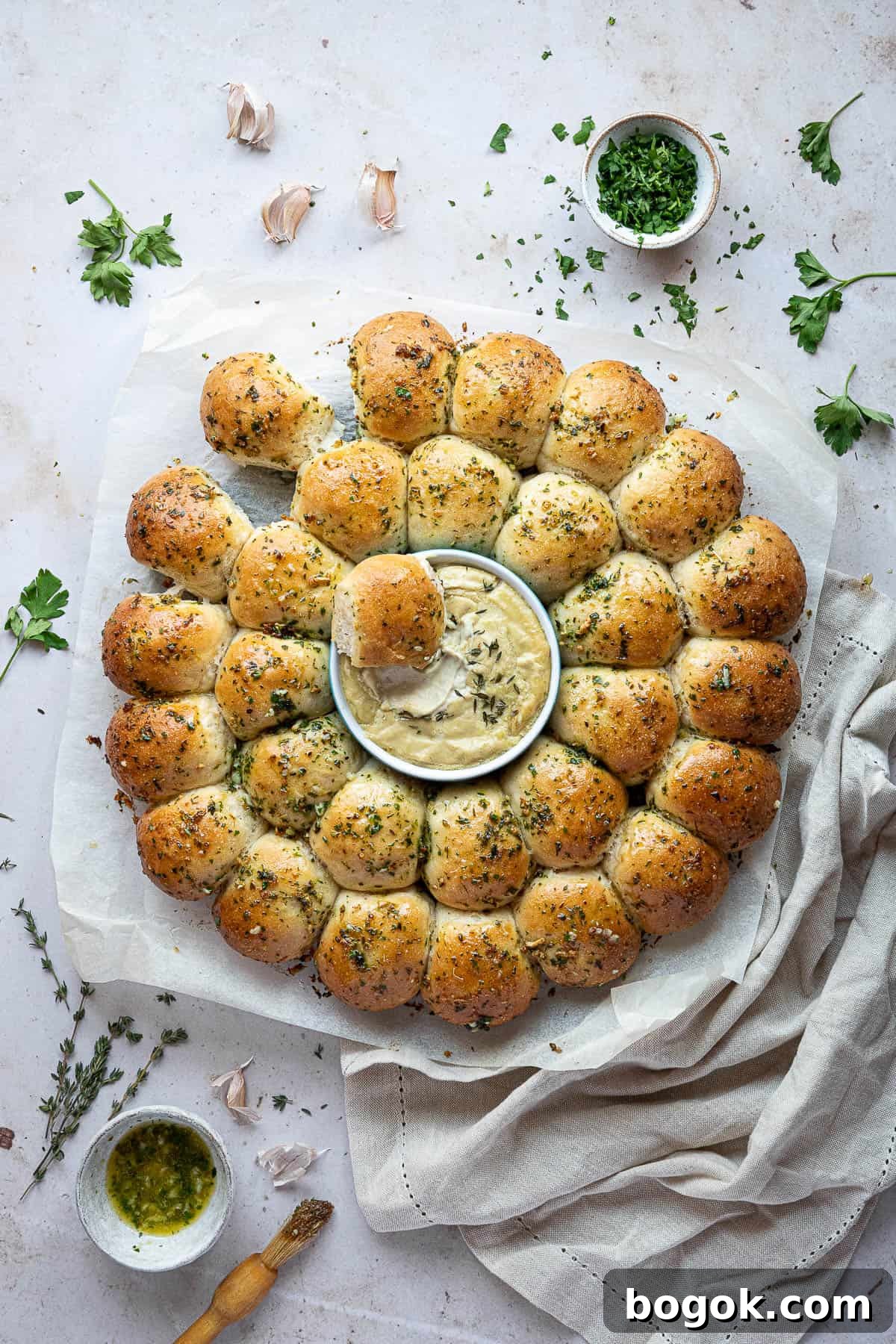 Vegan garlic dough balls partially torn apart on a sheet of baking parchment with bowls of garlic butter and herbs.