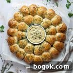 Vegan garlic dough ball wreath on a sheet of baking parchment surrounded by bowls of herbs and garlic butter.