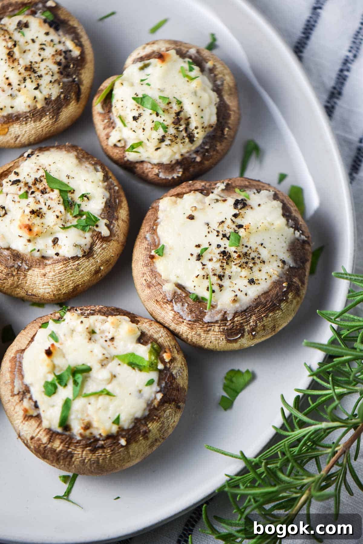 Boursin-Stuffed Mushrooms with Garlic and Herbs 9 Baked stuffed mushrooms topped with pepper and parsley next to rosemary leaves on a plate.