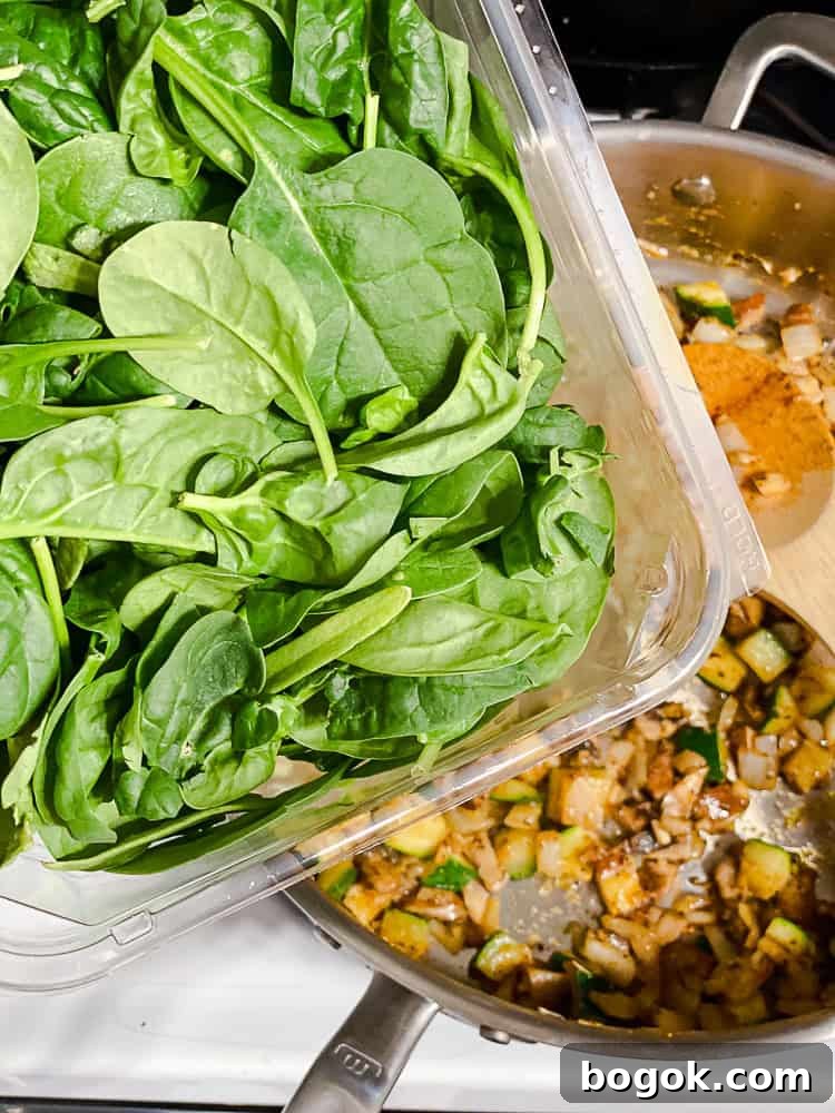 pouring spinach leaves into a skillet full of cooked vegetables.