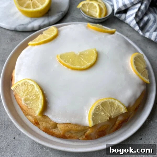 Round lemon cake with white icing, topped with six halved lemon slices, displayed on a white plate. A sliced lemon and cloth napkin are in the background.