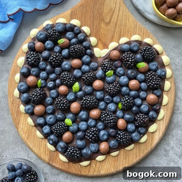 A heart-shaped wooden board topped with blueberries, blackberries, basil leaves, and chocolate candies on a layer of white and dark chocolate.