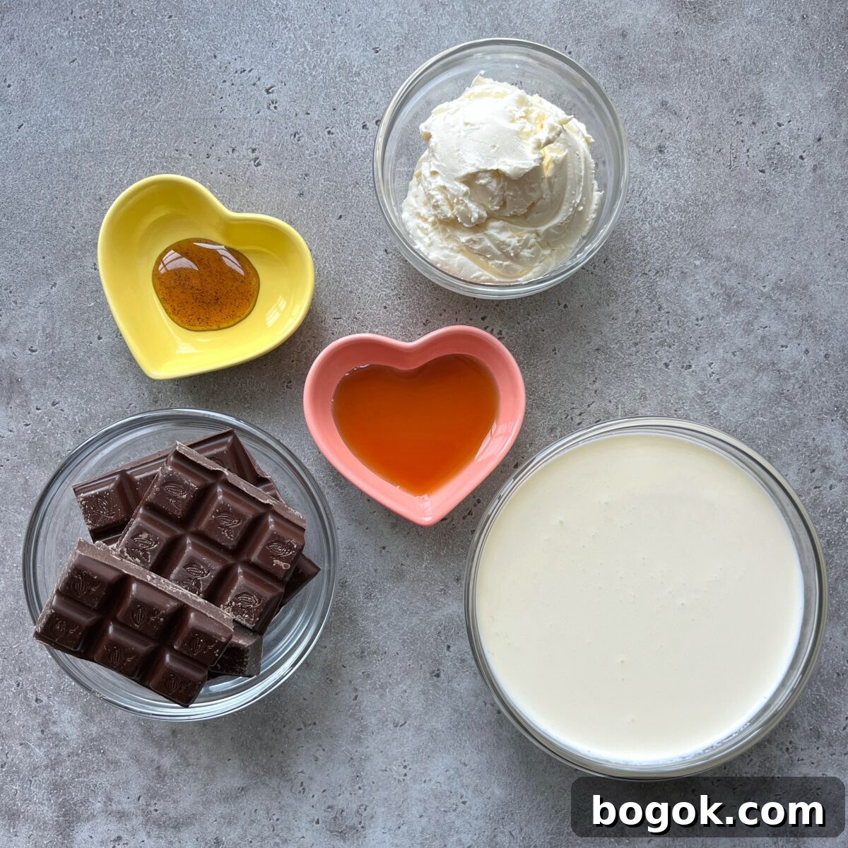 Five bowls containing chocolate pieces, heavy cream, mascarpone, maple syrup, and vanilla paste arranged on a gray surface.