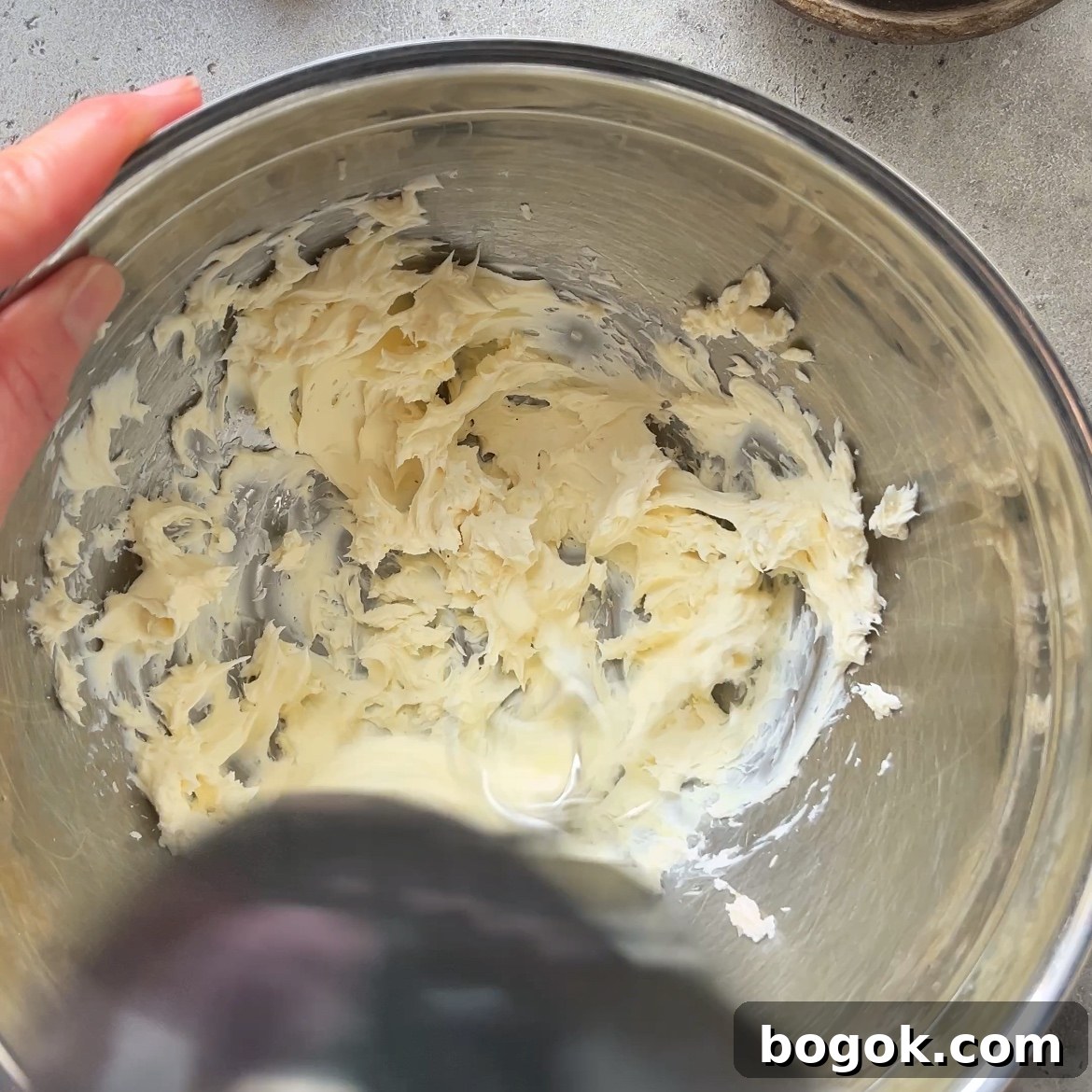 A hand holds a metal bowl with mascarpone and vanilla paste being whipped by an electric mixer on a gray countertop.