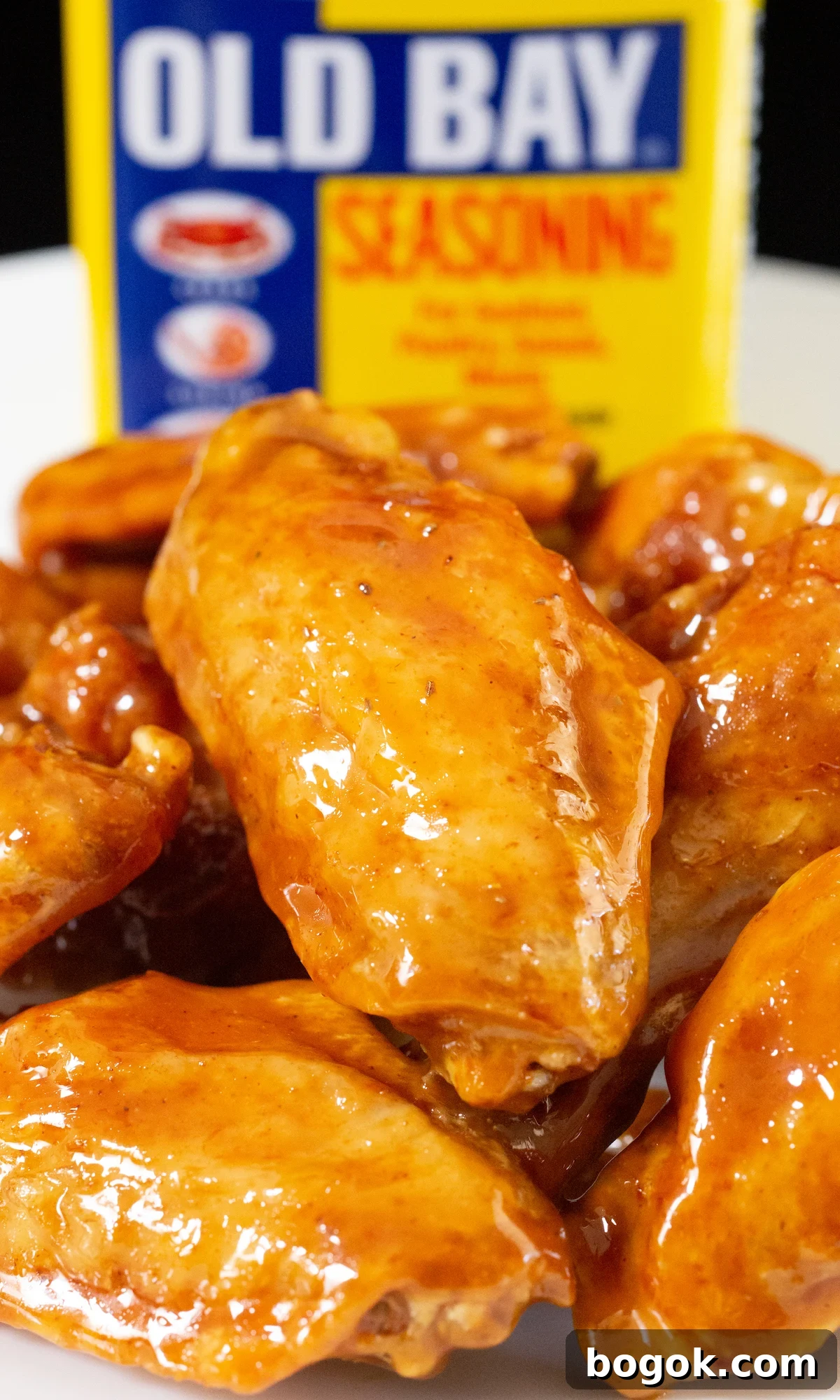 Close up of a flat chicken wing covered in a reddish-orange glaze. An Old Bay seasoning container is out of focus in the background.