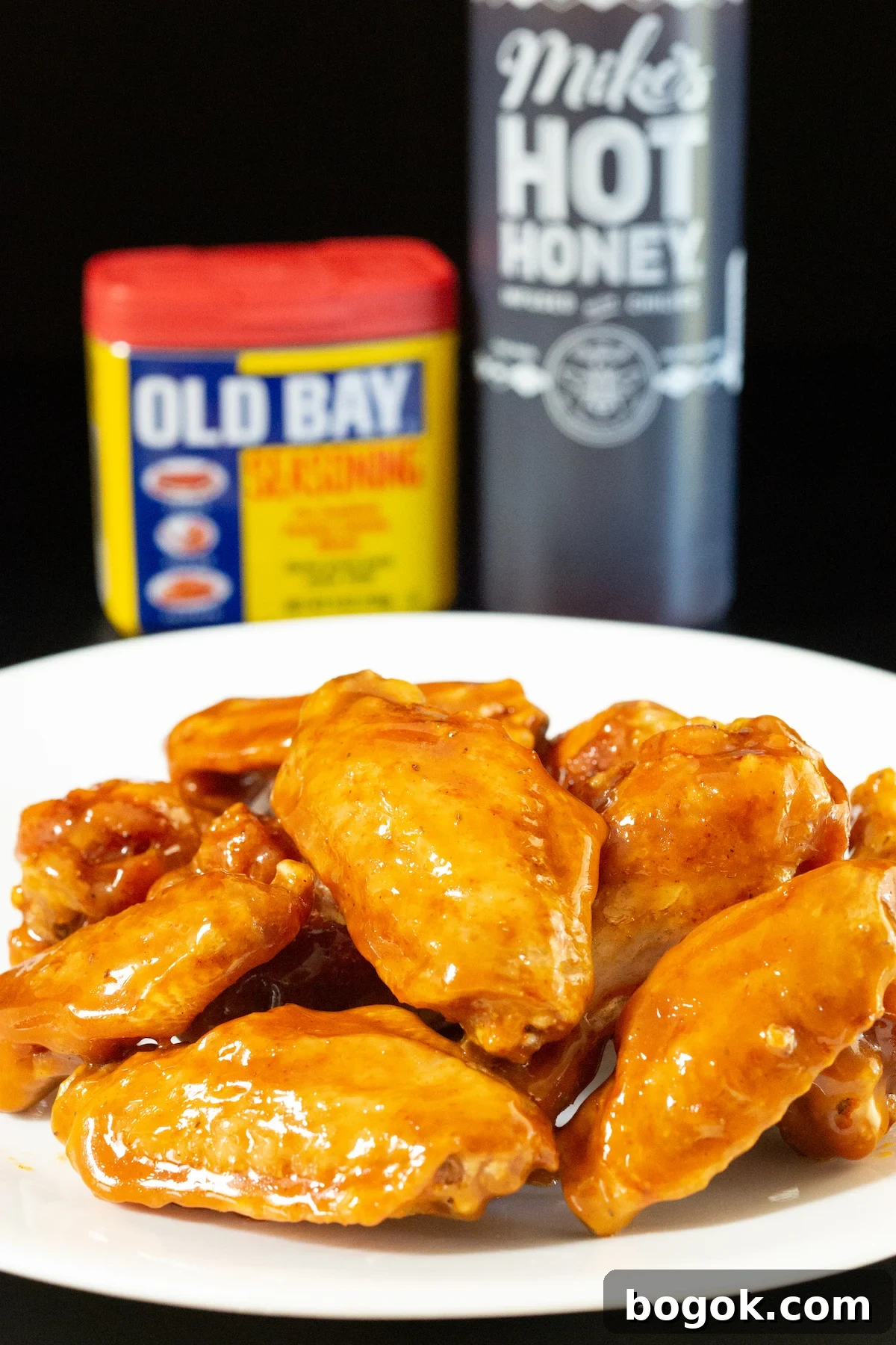 A dinner plate filled with about a dozen chicken wings covered in an orange colored glaze. An old bay seasoning container and a bottle of hot honey are out of focus in the background.