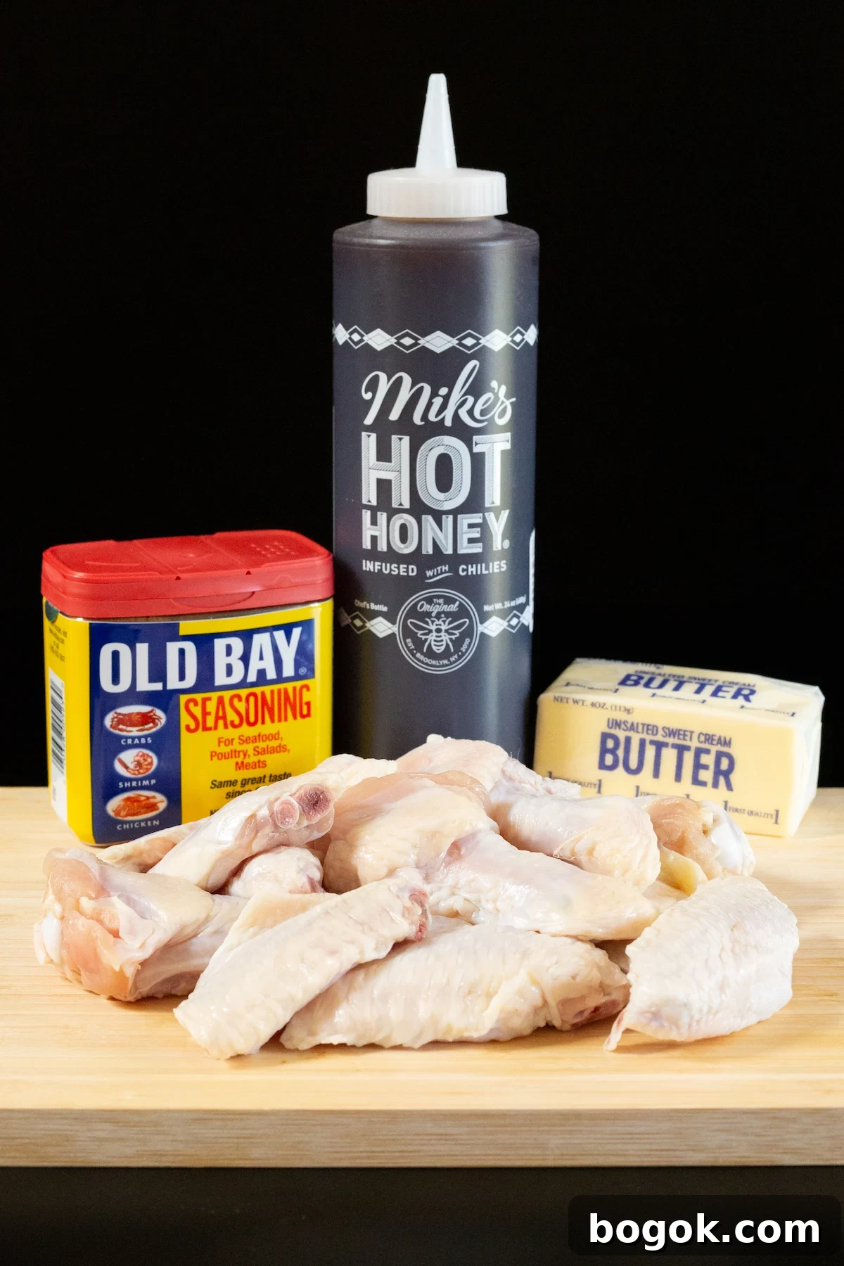 A bottle of Old Bay seasoning, Mike's Hot Honey, a stick of butter, and about a dozen chicken wings on a wood cutting board.