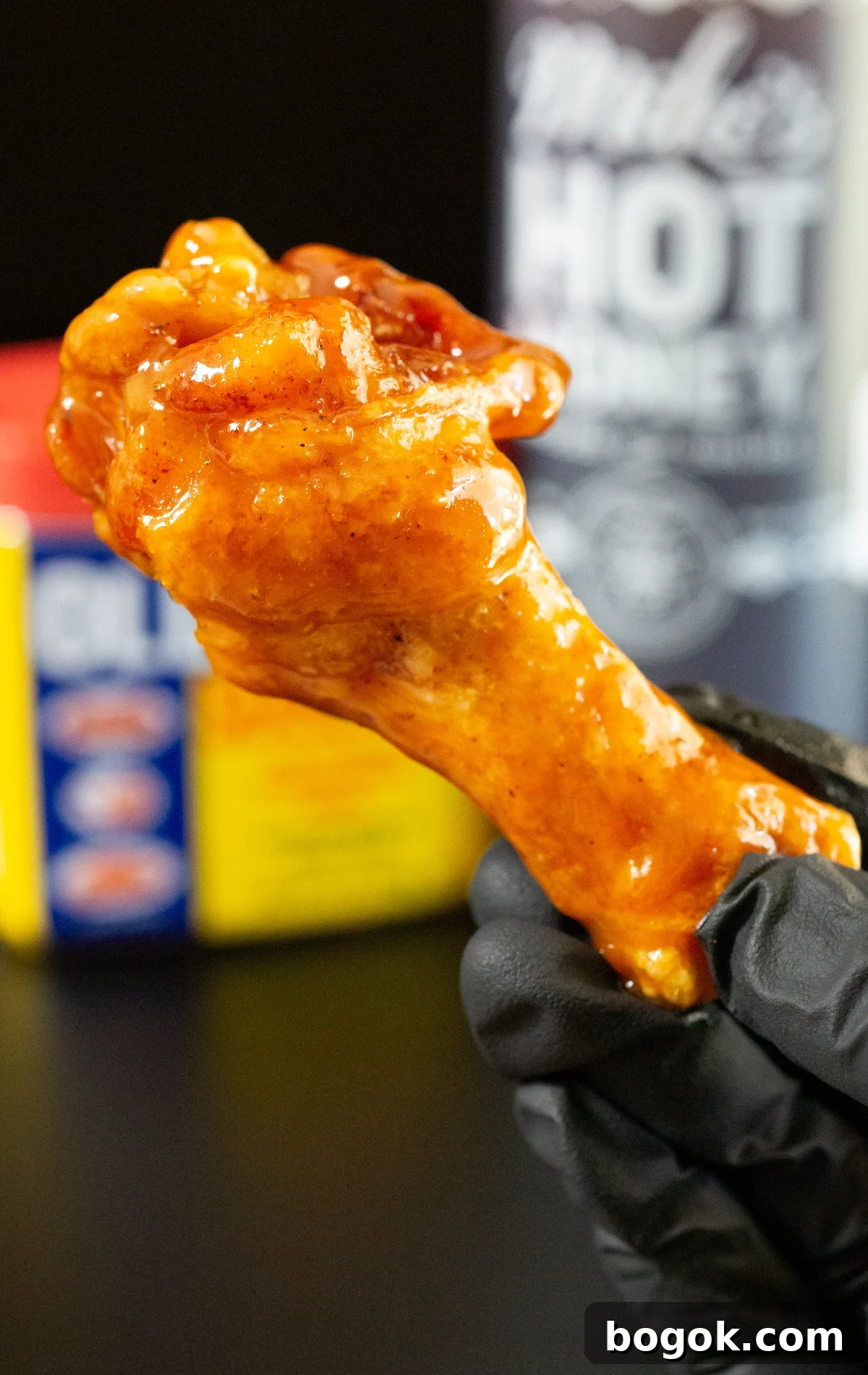 A hand olds up a drumstick chicken wing covered in a shiny glaze. In the background, a bottle of Mike's Hot Honey and and Old Bay seasoning are out of focus.