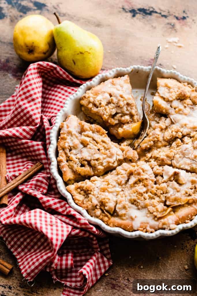 Sliced pear dessert pizza in a pie dish on a wooden surface surrounded by a red checked linen, cinnamon sticks, and fresh pears