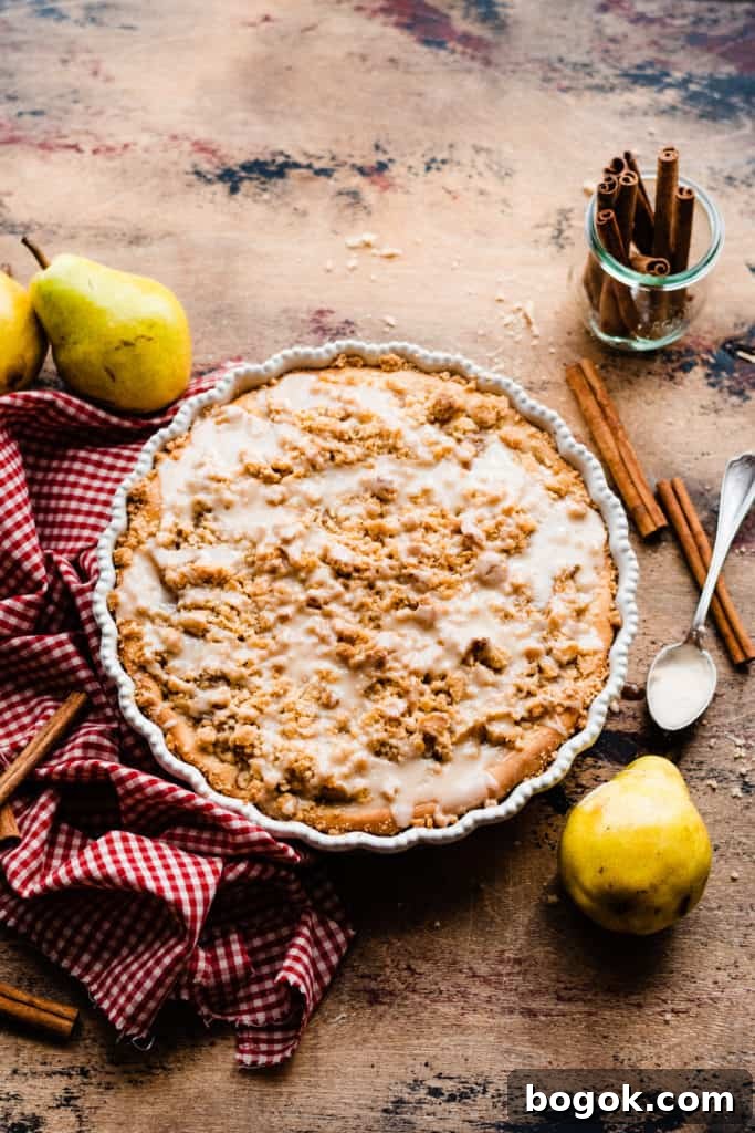 Pear dessert pizza baked and drizzled with glaze on a wooden surface surrounded by a red checked linen, cinnamon sticks, and fresh pears