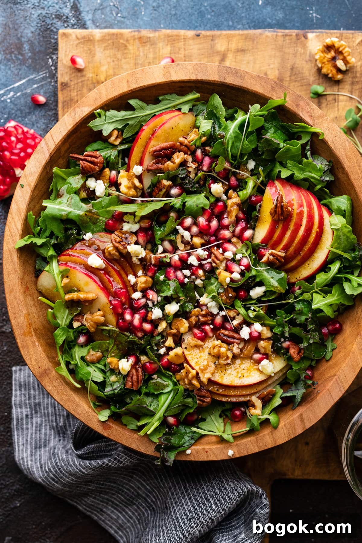 A wooden bowl with apple pomegranate salad and a bed of greens in it. The bowl of salad is sitting on a wooden cutting board with a dark background behind it. With pomegranate seeds and a napkin arranged around the salad.