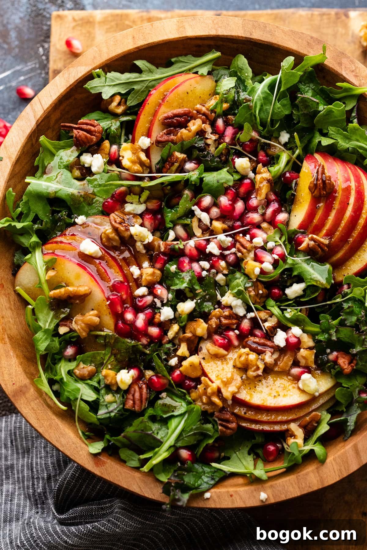 A close up of apple pomegranate salad in a bed of greens in a wooden bowl. 