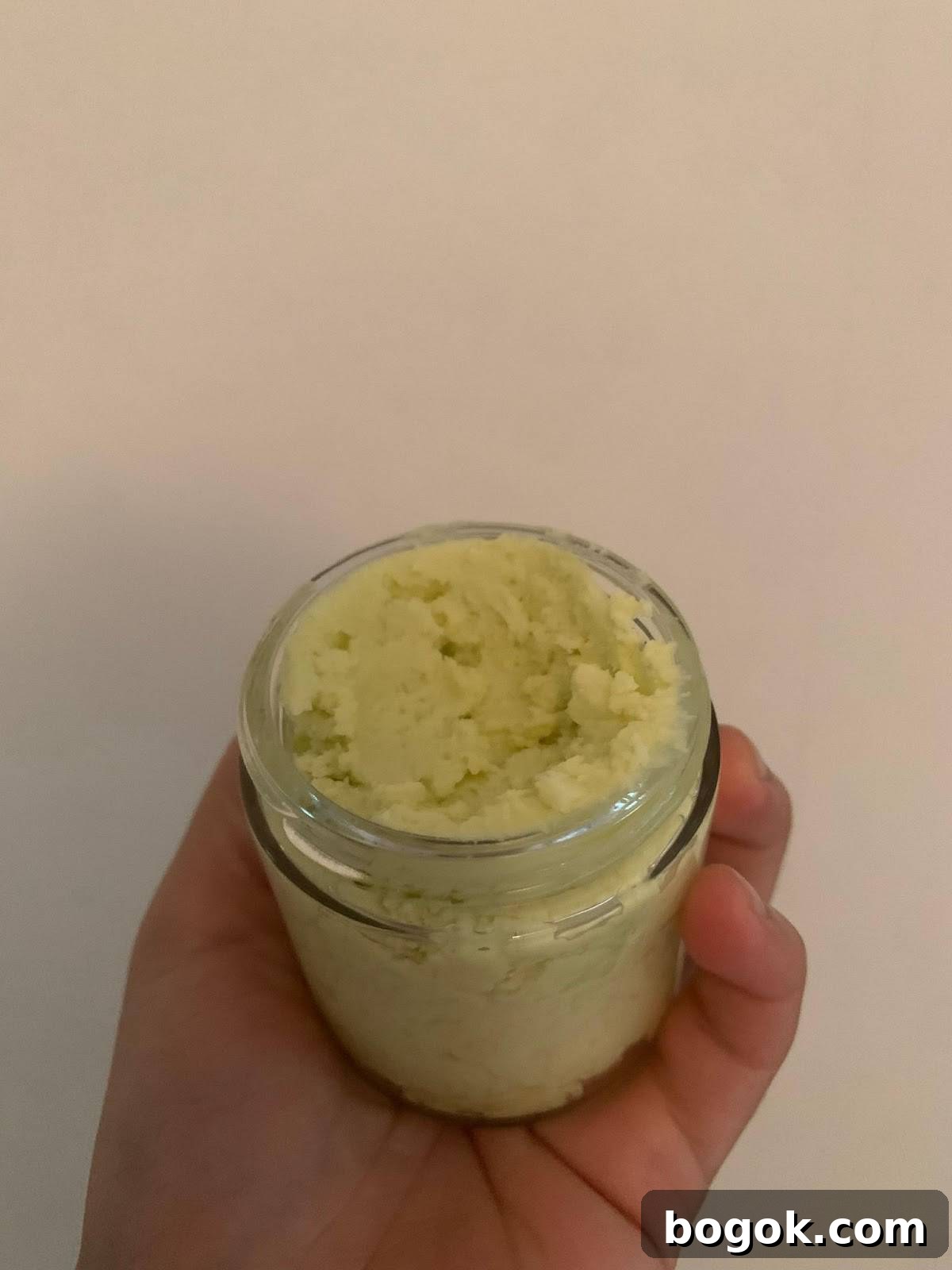 A hand is holding a small, clear glass jar filled with a calendula body lotion