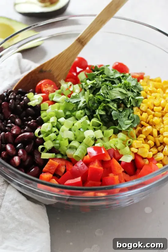 Healthy and easy southwestern bean salad! Packed with two kinds of beans, colorful veggies, creamy avocado and a honey lime chipotle dressing! The whole dish can be made in advance, making it perfect for meal prep, parties or picnics! Gluten and dairy free! A glass bowl filled with all the components for Southwestern Bean Salad with a wooden spoon in the dish.