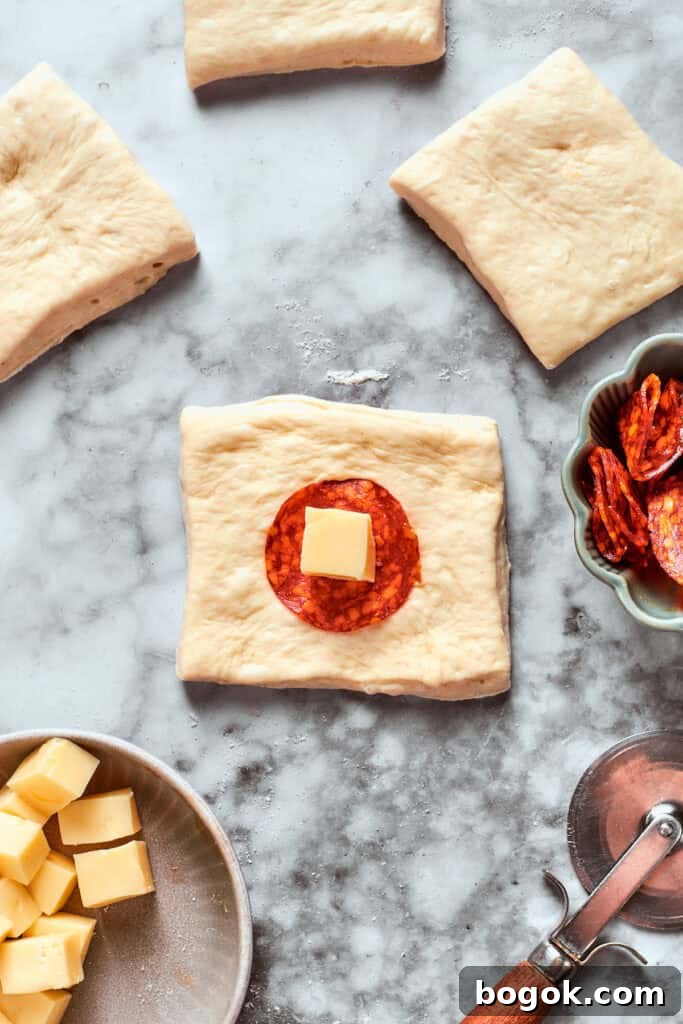 A slice of pepperoni and a cube of cheese is added to a square of dough.