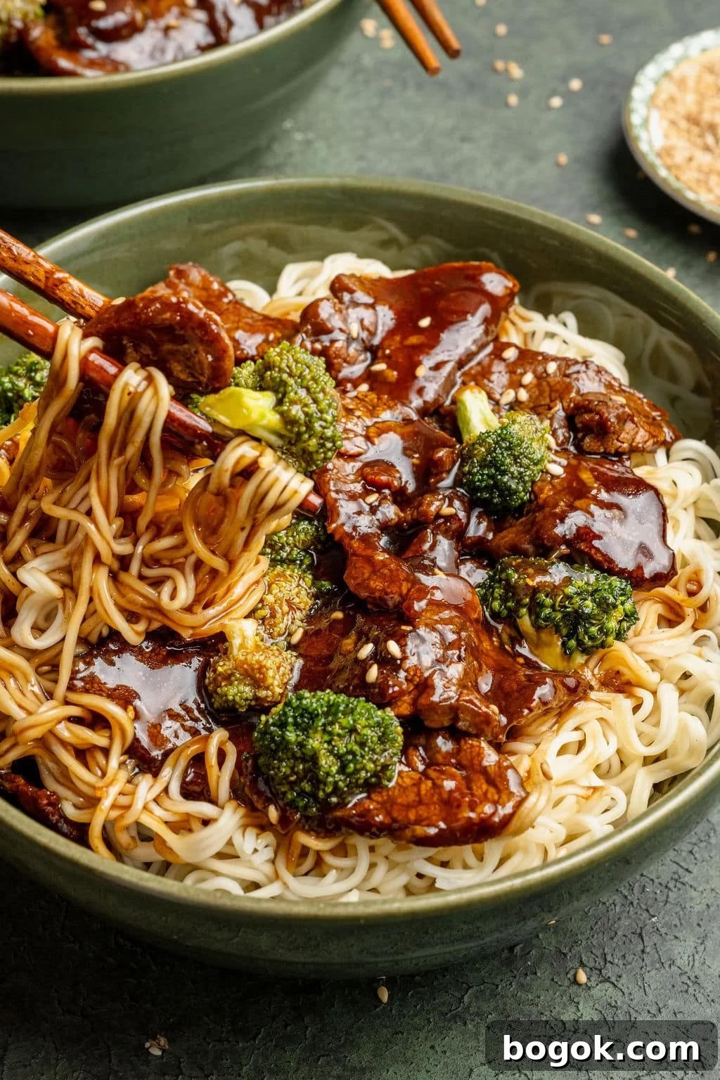 Tight shot of chopsticks lifting noodles from a bowl of beef and broccoli, highlighting the glossy sauce, sesame seeds, and tender pieces of beef.