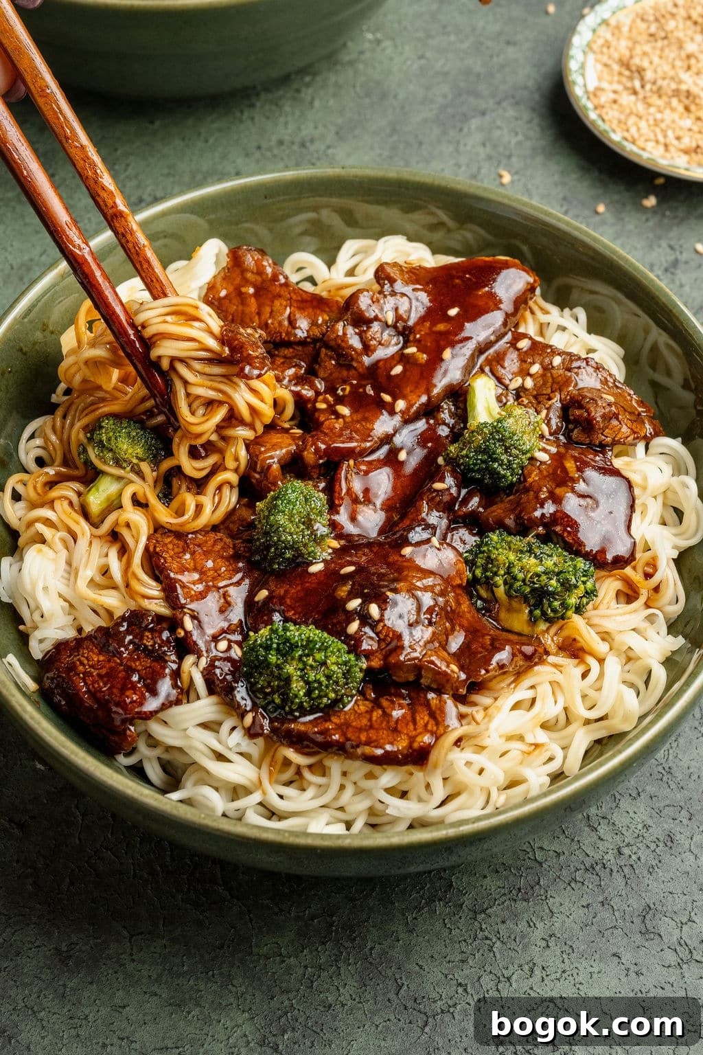 Close-up of beef and broccoli noodles with chopsticks lifting a bite of noodles coated in savory sauce.