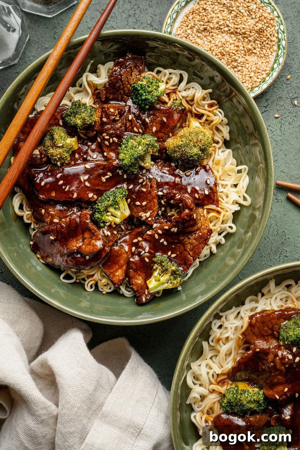 Top-down view of two green bowls filled with beef and broccoli over noodles, sprinkled with sesame seeds.