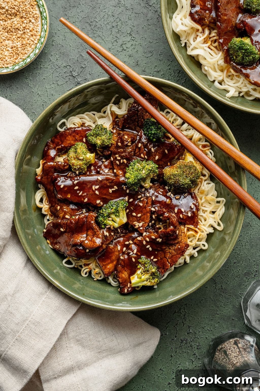 Bowl of beef and broccoli over ramen noodles, garnished with sesame seeds and served with chopsticks resting on the rim.