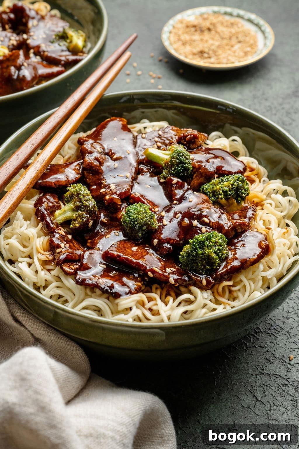 Side view of beef and broccoli noodles in a green bowl with chopsticks, showing glossy sauce and sesame seed garnish.