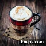 A large mug filled with a delicious homemade latte, topped with a sprinkle of cinnamon, with loose coffee beans on the table.