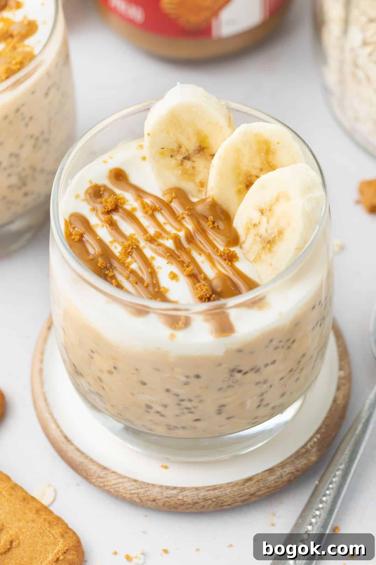 Creamy Biscoff Overnight Oats Recipe for Quick Breakfast 2 Glass jar of overnight oats topped with yoghurt, slices of banana and a drizzle of Biscoff spread, sitting on a round coaster.
