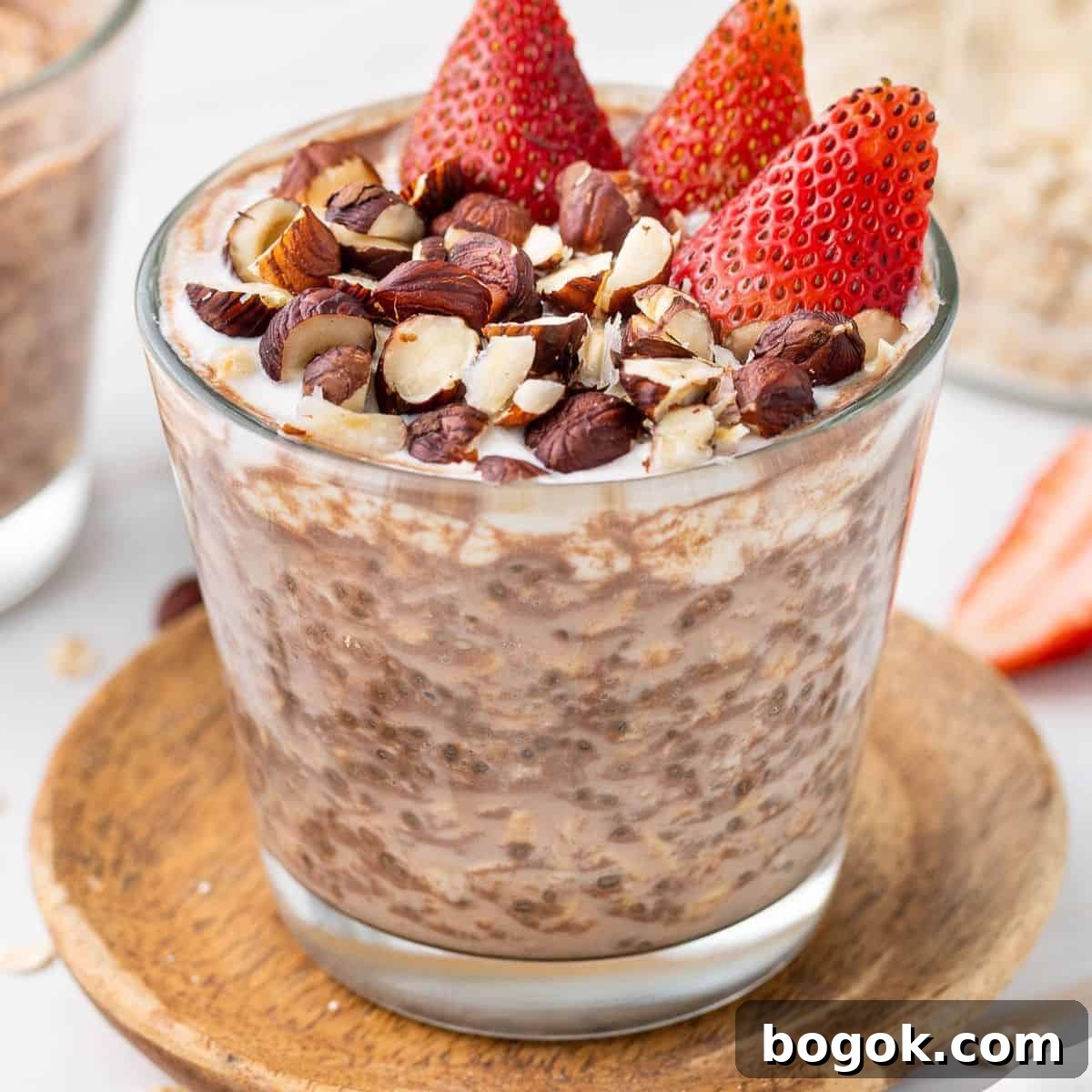 Glass jar of overnight oats, topped with yoghurt, crushed hazelnuts and cut strawberries, sitting on a round wooden coaster.