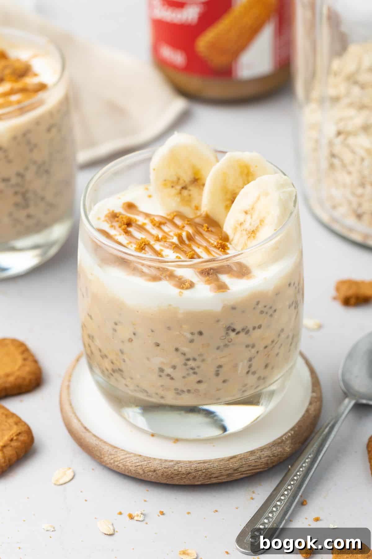 Creamy Biscoff Overnight Oats Recipe for Quick Breakfast 6 Glass jar of overnight oats topped with yoghurt, slices of banana and a drizzle of Biscoff spread, sitting on a round coaster.