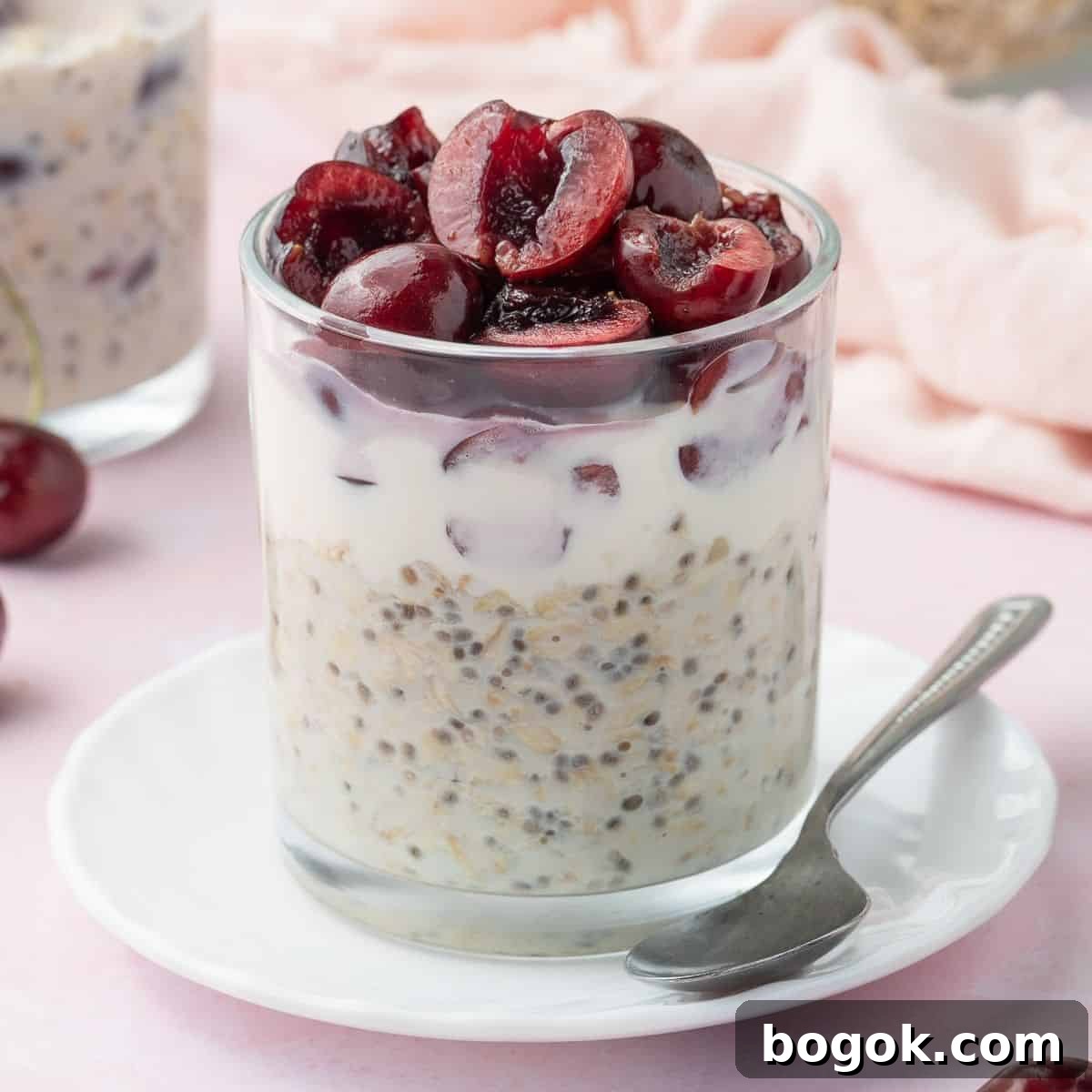 Glass jar of vanilla overnight oats, topped with halved cherries, sitting on a small white saucer with a teaspoon on the edge.