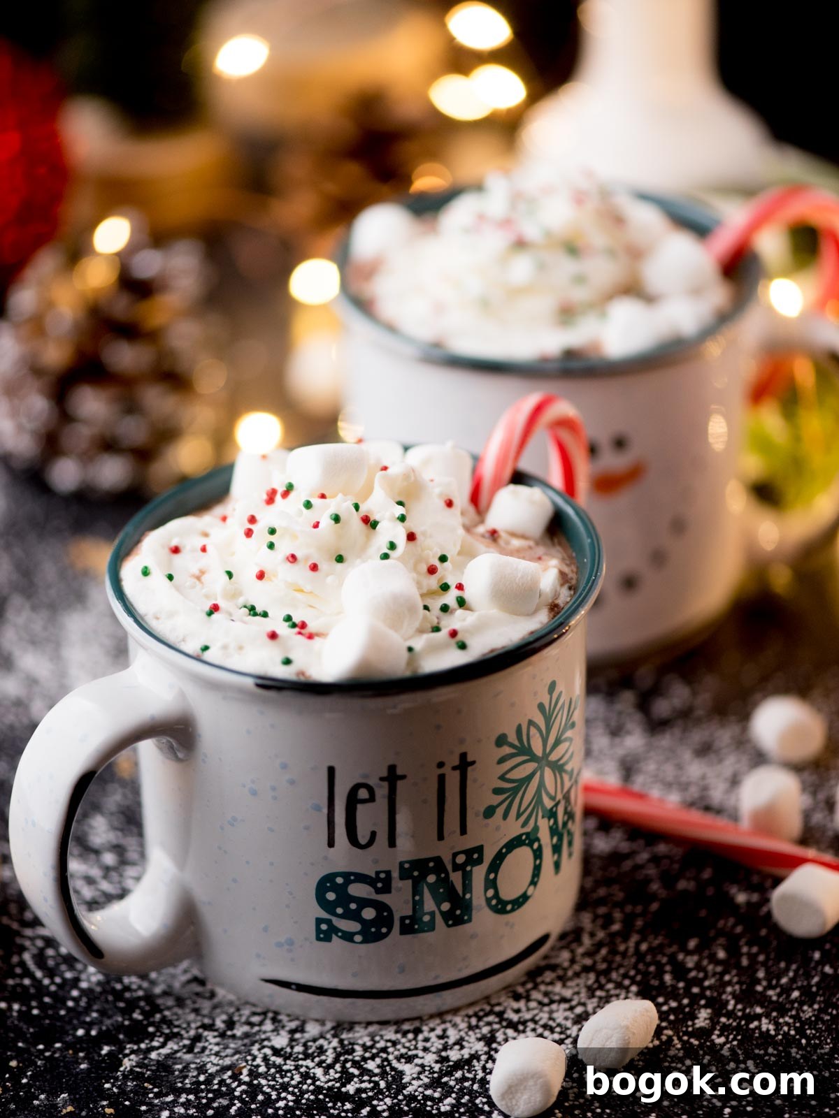 Two mugs of hot chocolate topped with whipped cream, colorful sprinkles, marshmallows, and candy canes. The front mug features the text let it snow. A festive background with fairy lights and snow-like powder completes the scene.