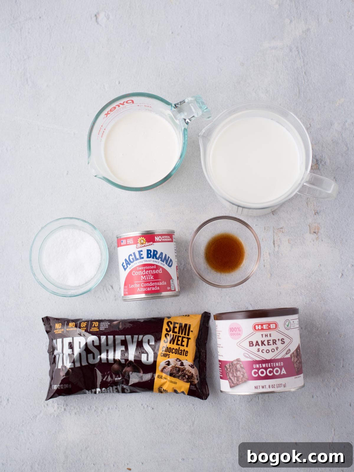 Ingredients for a dessert are laid out on a light surface: a bag of semi-sweet chocolate chips, a can of sweetened condensed milk, a small bowl of sugar, a glass of vanilla extract, a carton of heavy cream, and a box of unsweetened cocoa.
