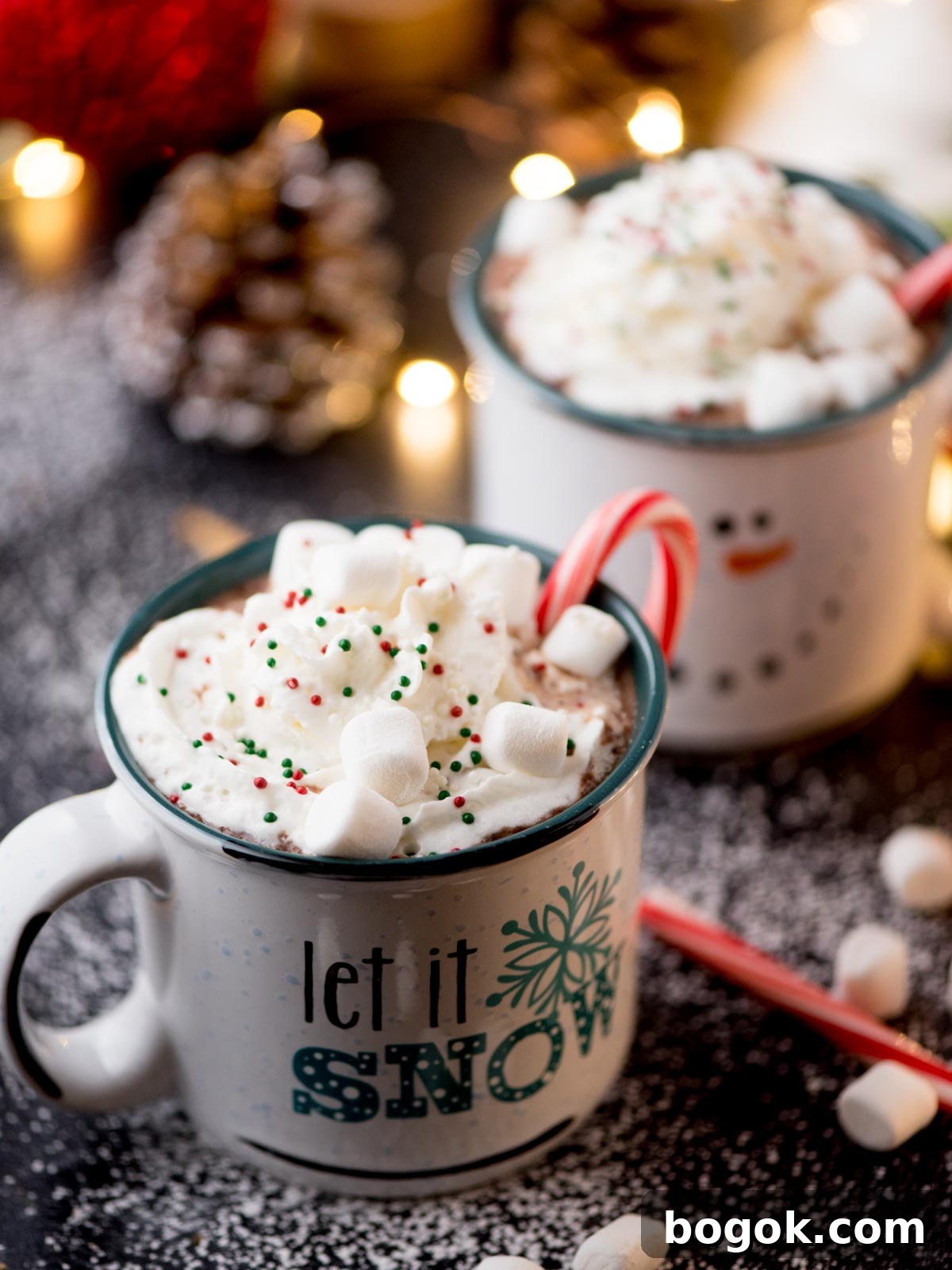 Two festive mugs of hot chocolate topped with whipped cream, sprinkles, and marshmallows. One mug has a candy cane and Let it Snow printed on it. The scene is sprinkled with powdered sugar, and there are twinkling lights and a pinecone in the background.